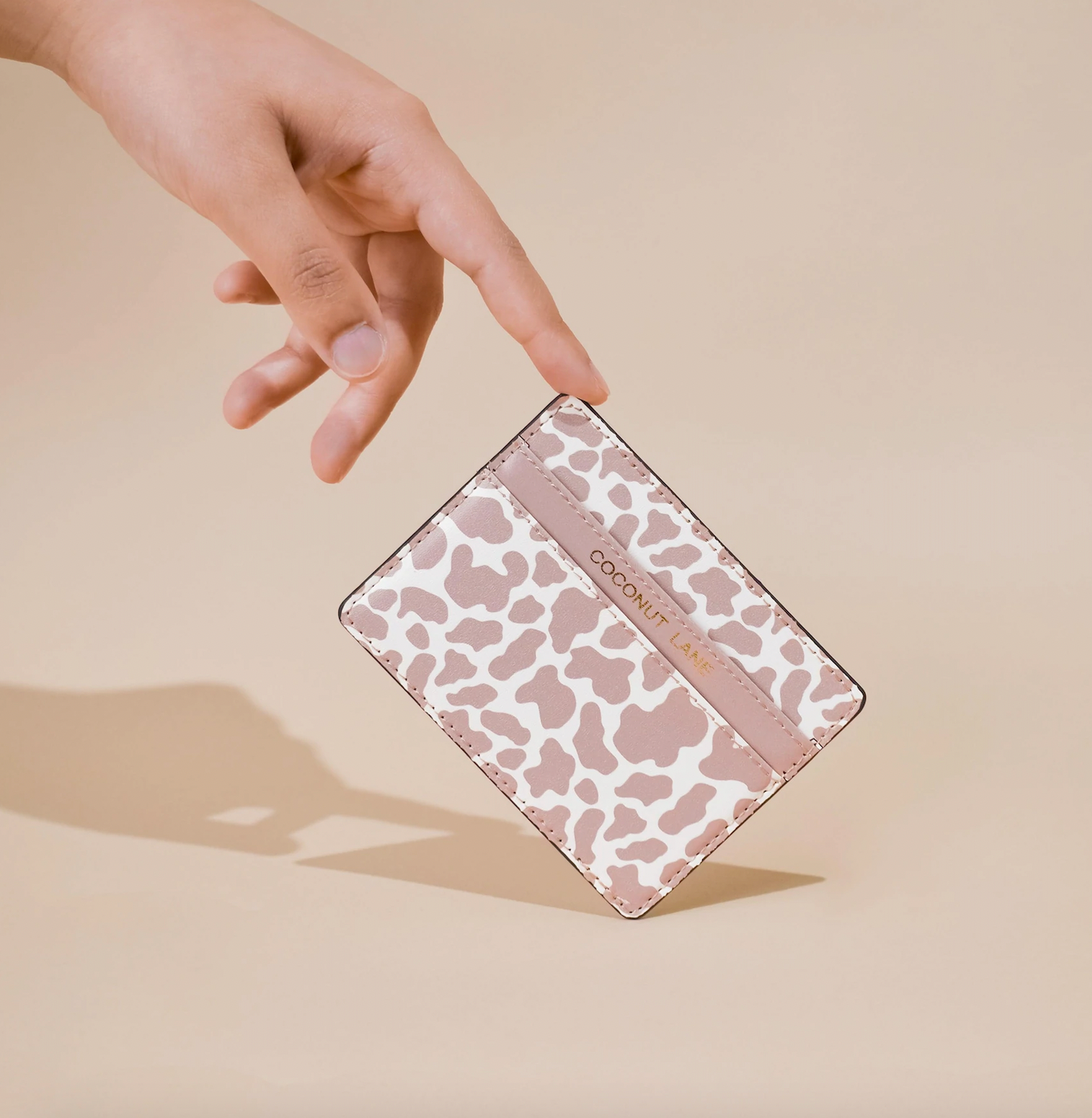 image of a card wallet with 2 sections. the middle section has the word coconut lane embossed in gold foil on it. the print is similar to a cow print but it is in pale pink and white