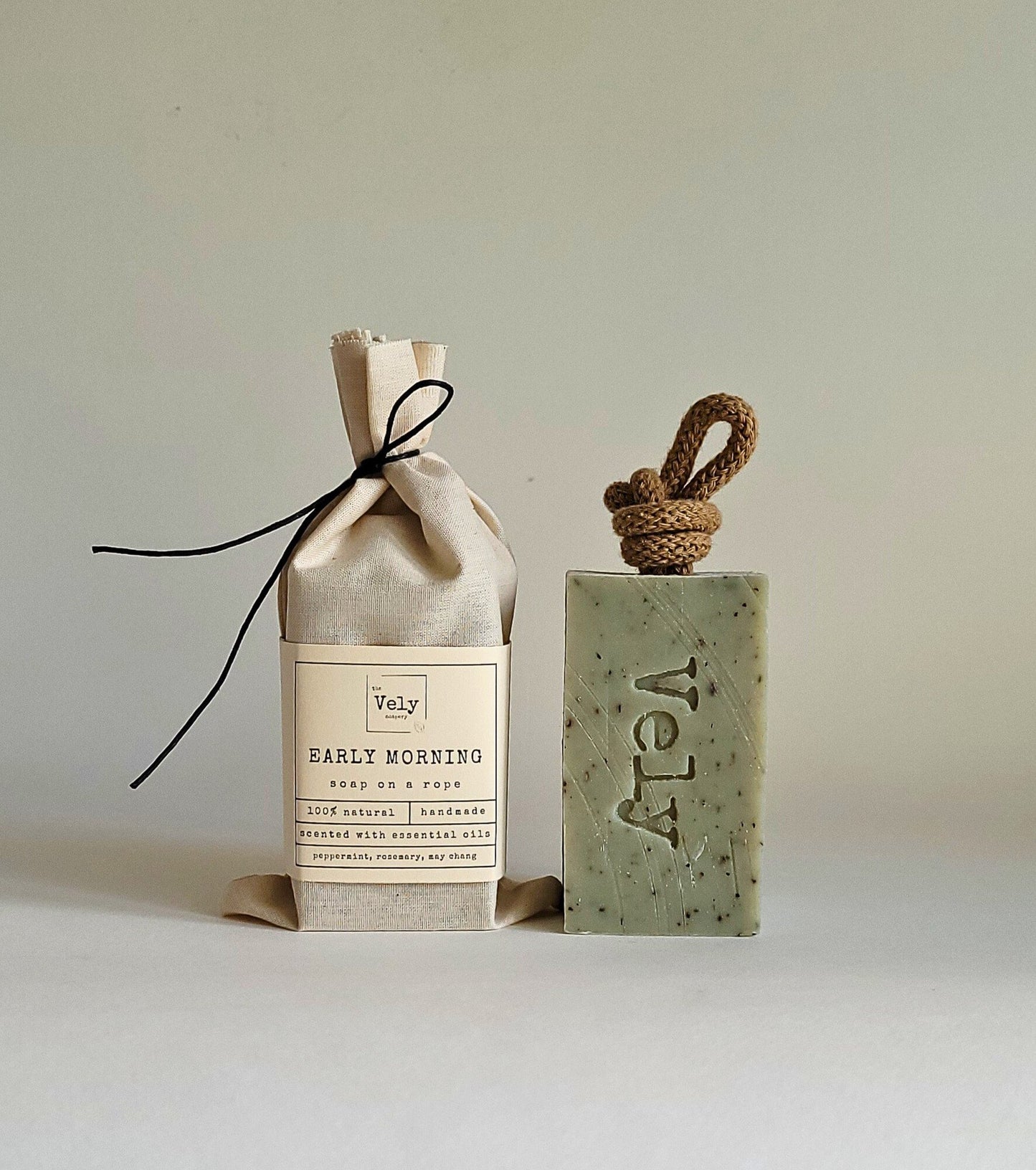 Green soap bar with brown rope. the word Vely is etched into the bar. The fragrance is early morning. The hessian bag that the soap comes in is shown to the left of the soap.