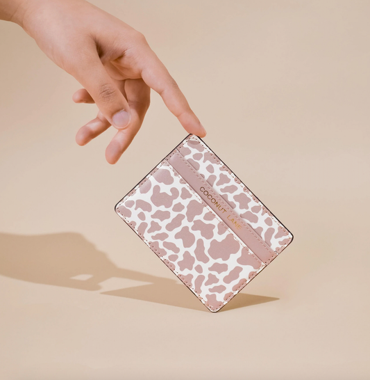 image of a card wallet with 2 sections. the middle section has the word coconut lane embossed in gold foil on it. the print is similar to a cow print but it is in pale  pink and white