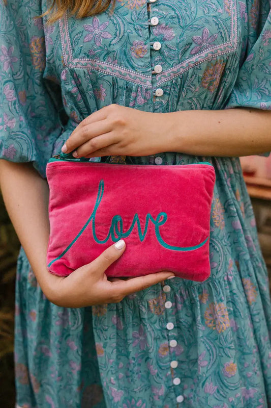 medium size pouch in pink velvet type material with the word love embroidered across the front in turquoise thread. There is a turquoise tassel on the zip.