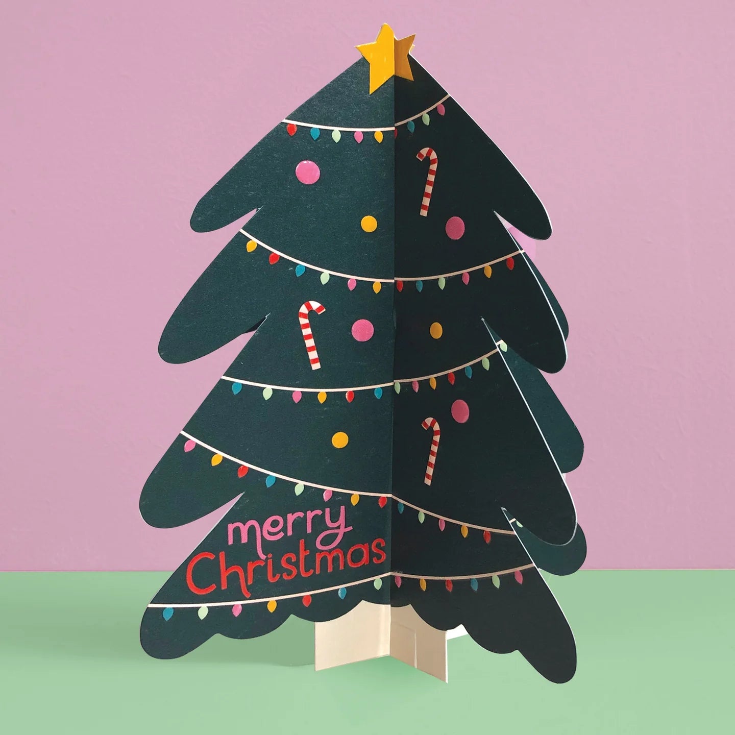 A 3D pop-up cardboard Christmas tree card on a light beige stand, set against a split pink and mint green background. The tree is dark green with a gold star on top, decorated with colorful string lights, candy canes, and the text 'merry Christmas' in pink and red.