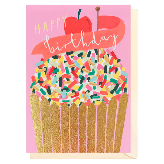 a colourful birthday card featuring a large cupcake with a golden wrapper. Above the cupcake are a cherry and pink banner stating happy birthday. Background is pink. The card blank inside and has a cream envelope