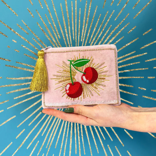 Pink pouch with cherry design held by a hand against a blue background with gold accents