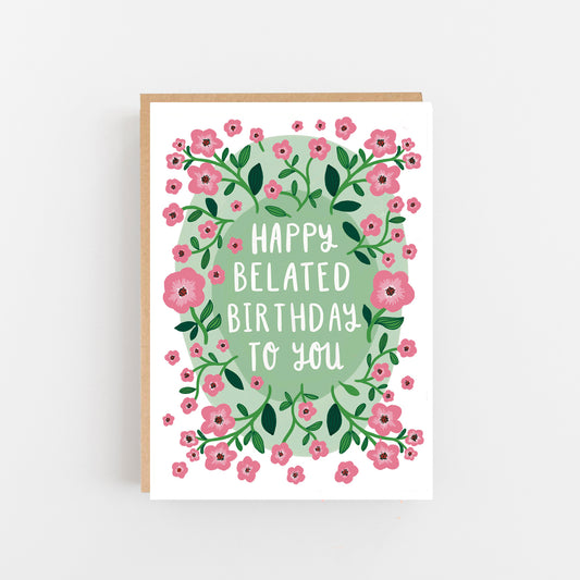 Greeting Card with the words Happy Belated Birthday to you written in White Capitals in the centre of the card on a pale blue background and surrounded by the drawings of pale pink and green flowers.