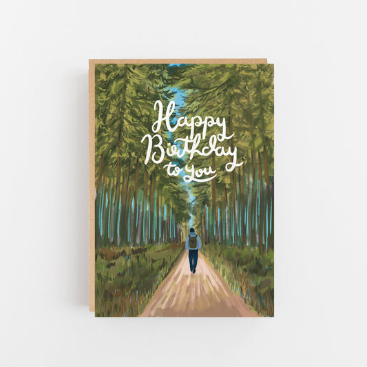 Greeting Card with the words Happy Birthday to you in white swirl writing in the centre front of the card. The background of the card is a drawing of a forest and the outline of a man dressed in pale blue and dark blue carrying a rucksack waling down the centre of a path between the trees.