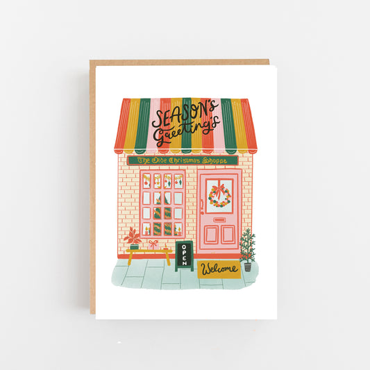 Christmas Card with a white background and a drawing of a shop front with a multi coloured stripped roof with the words Seasons greetings in black italics across the roof, The shop is mainly pale pink with a Christmas tree in the shop window and a wreath on the door. Outside the shop a Welcome Mat a n opening A Board sign a small table with presents on and a small Christmas tree by the door.