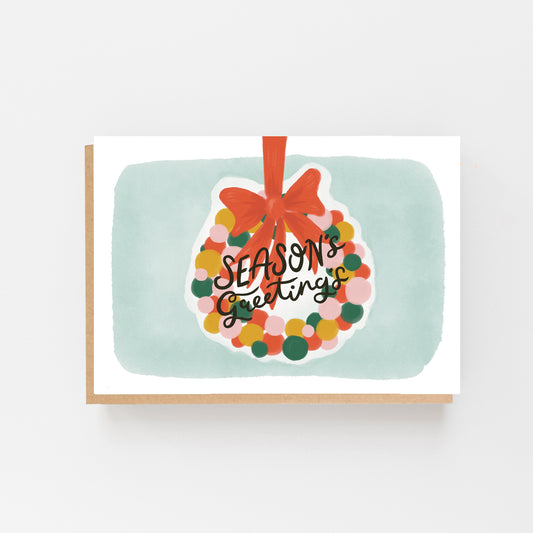 Christmas Greeting Card in landscape with a white and pale blue background. In the centre is the drawing of a wreath made up of pastel coloured circles and a big red bow at the top with the words Seasons Greetings written in Black Italics across the centre of the wreath. Brown Kraft Envelope.