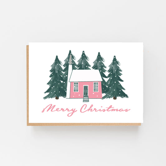 Christmas Card in landscape with a white background with green forest pine trees in the background with snow falling in the centre the drawing of a house in pale pink and white . The words Merry Christmas written in Pale pink italics at the bottom of the house. Brow Kraft Envelope.