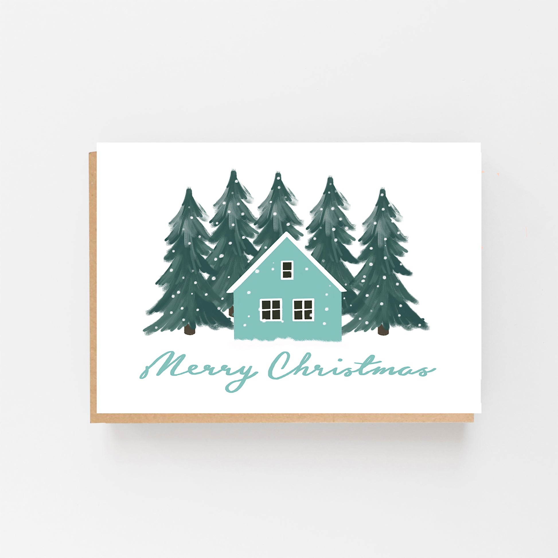 Christmas Card in landscape with a white background with green forest pine trees in the background with snow falling in the centre the drawing of a house in pale blue and white . The words Merry Christmas written in Pale blue italics at the bottom of the house. Brown Kraft Envelope