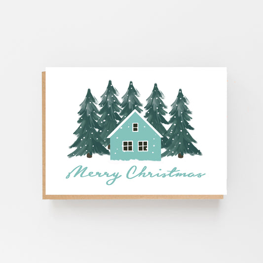 Christmas Card in landscape with a white background with green forest pine trees in the background with snow falling in the centre the drawing of a house in pale blue and white . The words Merry Christmas written in Pale blue italics at the bottom of the house. Brown Kraft Envelope