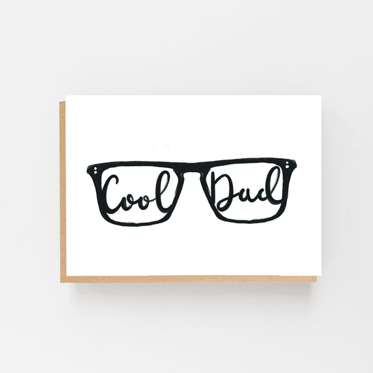 Greeting Card in landscape with the drawing of a pair of glasses in black on a white background with the words Cool in one lense and Dad in the other in Black Italic writing. Brown Kraft Envelope.