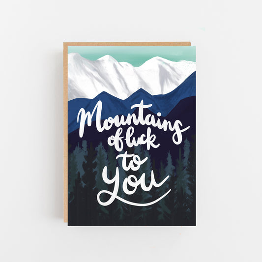 Greeting Card with the words Mountains of Luck to you written in white italics in the centre the background is a drawing of trees and mountains in dark blue green and white. Brown Kraft Envelope.