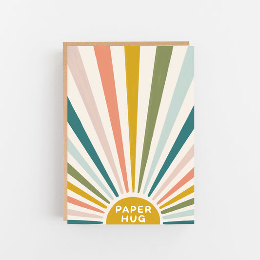 Greeting Card with the words Paper Hug in white writing in capital letters on a mustard coloured small half moon shape with rays in multi coloured pastel  colours looks like the sun's rays . Brown Kraft Envelope