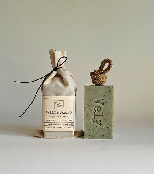 Green soap bar with brown rope. the word Vely is etched into the bar. The fragrance is early morning. The hessian bag that the soap comes in is shown to the left of the soap.