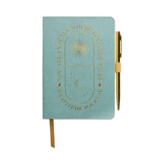 image shows an a5 notebook in pale blue with gold embossed words that are written around a long oval shape. the words read you are exactly where you need to be then a sun mofit and the word gratitude journal. Placed on the righthand side of the book is an elastic loop with a gold ball point pen inside the loop
