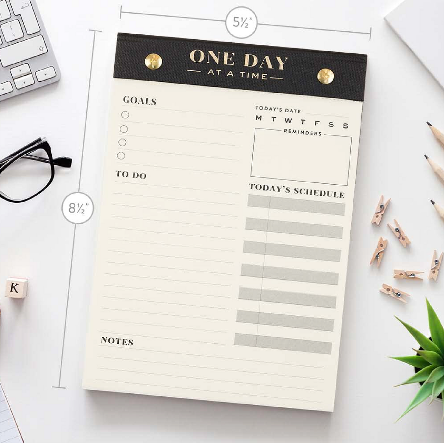 A daily planner notepad titled 'ONE DAY AT A TIME' laid flat on a white desk with measurement callouts showing 5 1/2 inches wide and 8 1/2 inches tall. The pad has a black top band with gold rivets and gold lettering, cream pages, lined sections for goals, to-do, schedule, and notes, and a date/reminder box.