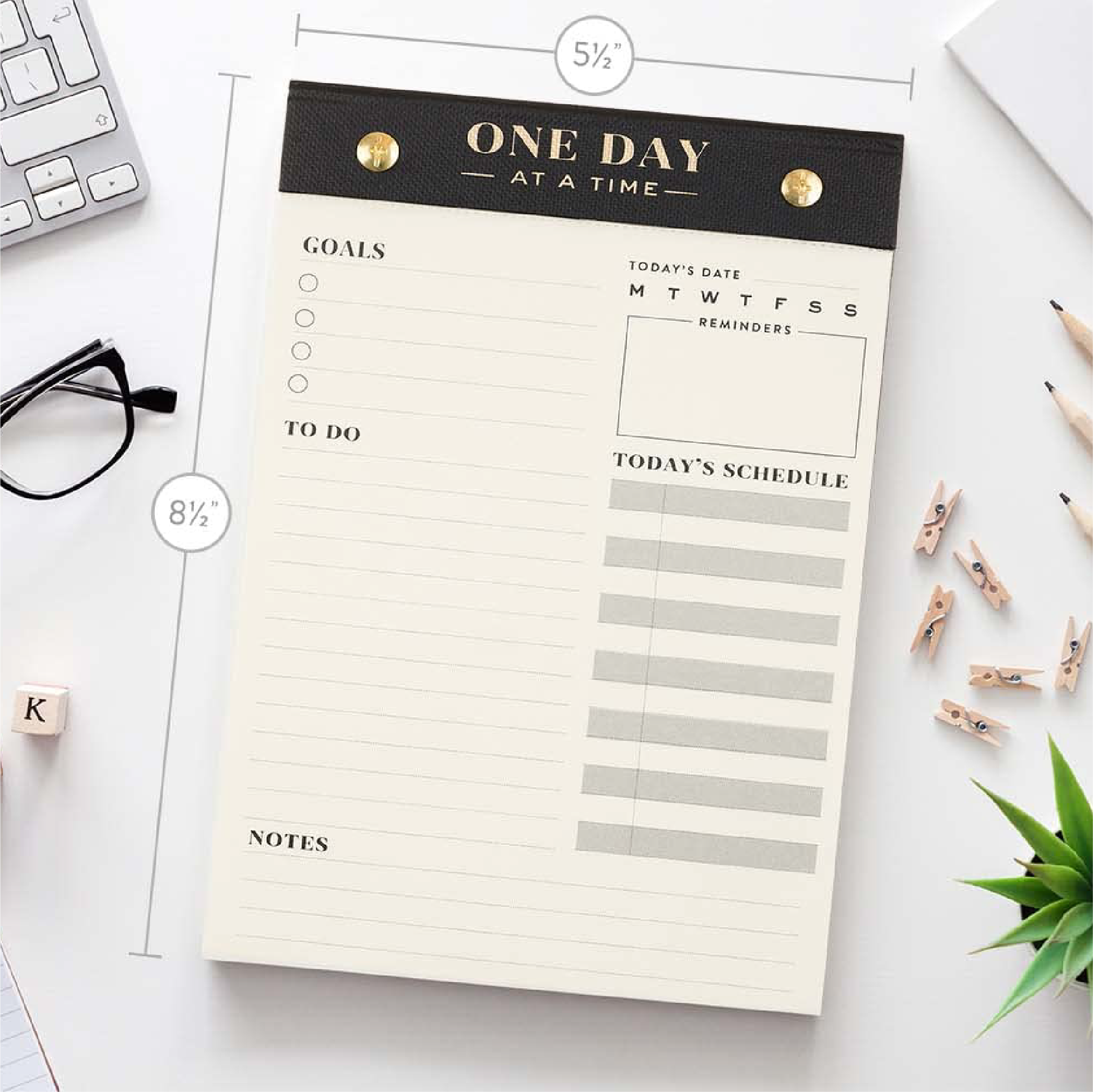 A daily planner notepad titled 'ONE DAY AT A TIME' laid flat on a white desk with measurement callouts showing 5 1/2 inches wide and 8 1/2 inches tall. The pad has a black top band with gold rivets and gold lettering, cream pages, lined sections for goals, to-do, schedule, and notes, and a date/reminder box.
